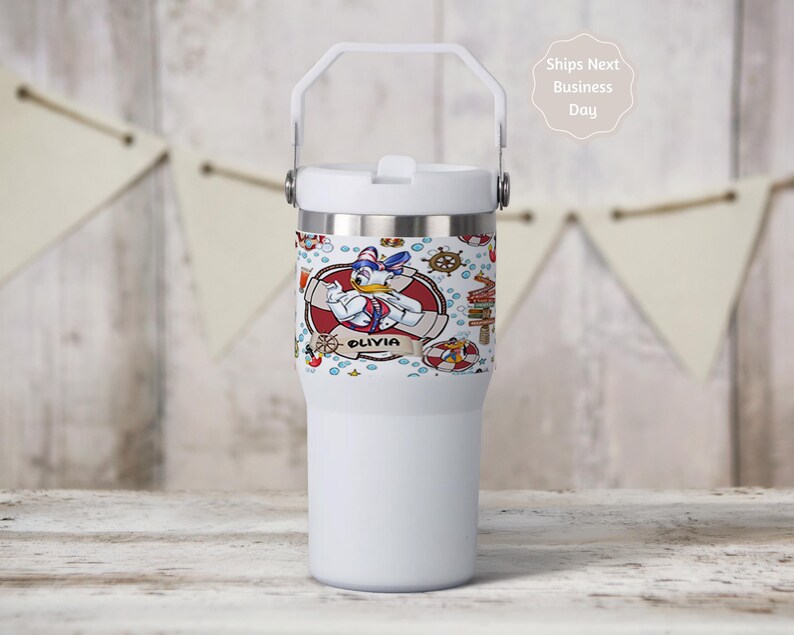 May include: A white insulated tumbler with a handle and a lid. The tumbler features a wrap-around design with a cartoon Donald Duck illustration, nautical elements, and the name "Olivia". The tumbler is designed to keep beverages cold or hot.