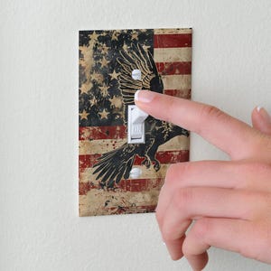 May include: A rectangular light switch wall plate featuring a distressed American flag design with a black eagle silhouette overlaid on the stars and stripes. The plate has a white toggle switch in the center with the word ON printed on it.