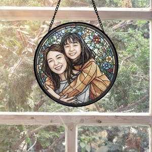 May include: A round, stained-glass window art piece featuring two smiling women embracing. The artwork is set against a backdrop of colorful floral patterns. The piece is framed in black and suspended by a chain, creating a decorative element.
