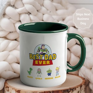May include: A white ceramic mug with a dark green interior and handle. The front features a graphic of Buzz Lightyear above the text "BEST DAD EVER" and four small character illustrations with the names Matt, Emily, Georgie, and Dylan below.
