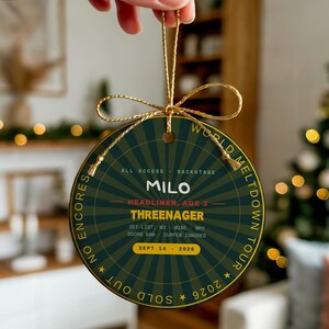 May include: A round, dark green ornament with a gold sunburst pattern and a gold string. Text reads: MILO, HEADLINER, AGE 3, THREENAGER, ALL ACCESS, BACKSTAGE, SET LIST: NO, MINE, WHY, DOORS 6AM, CURFEW IGNORED, SEPT 14, 2026, WORLD MELTDOWN TOUR, SOLD OUT.