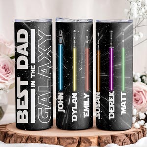K&ouml;nnte beinhalten: Drei schwarze isolierte Becher mit einem Galaxie-Design und dem Text "BEST DAD IN THE GALAXY". Jeder Becher hat ein unterschiedlich farbiges Lichtschwert und einen Namen: John, Dylan, Emily, Susan, Derek und Matt.
