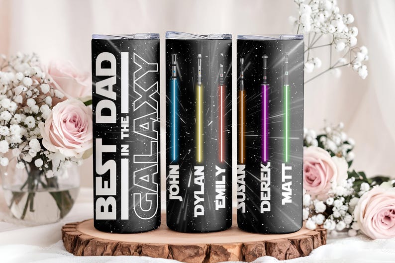 K&ouml;nnte beinhalten: Drei schwarze isolierte Becher mit einem Galaxie-Design und dem Text "BEST DAD IN THE GALAXY". Jeder Becher hat ein unterschiedlich farbiges Lichtschwert und einen Namen: John, Dylan, Emily, Susan, Derek und Matt.