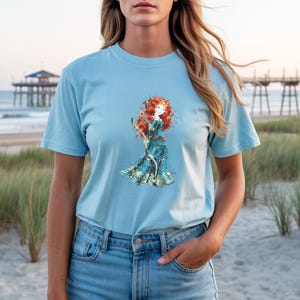 May include: Light blue t-shirt featuring a watercolor illustration of a red-haired character in a teal dress holding a bow and arrow. The shirt is made of soft material. The background shows a beach scene with a pier.