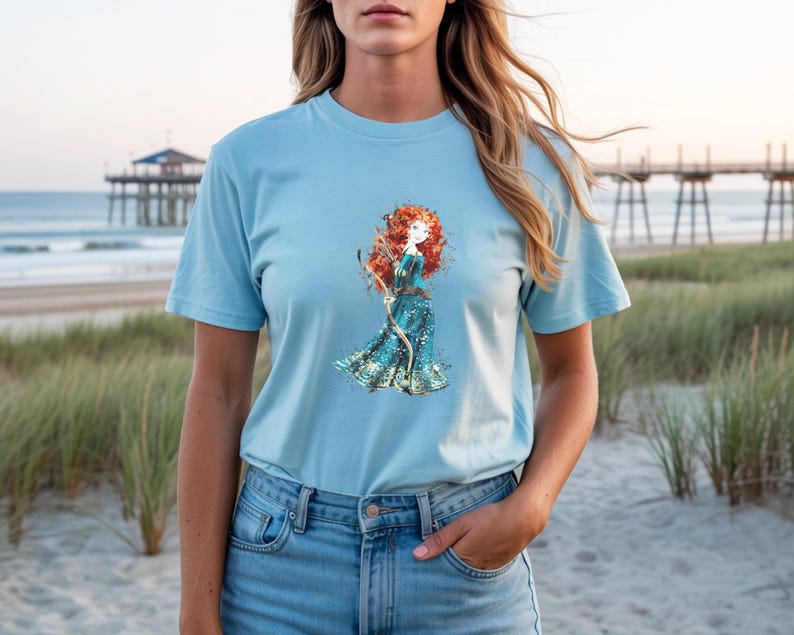 May include: Light blue t-shirt featuring a watercolor illustration of a red-haired character in a teal dress holding a bow and arrow. The shirt is made of soft material. The background shows a beach scene with a pier.