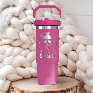 Puede incluir: Un vaso t&eacute;rmico rosa con asa y tapa a juego. El vaso presenta un gr&aacute;fico plateado de una mujer con gafas de sol y sombrero, con el texto "Squad" y el a&ntilde;o "2026". El vaso est&aacute; sobre una superficie de madera.