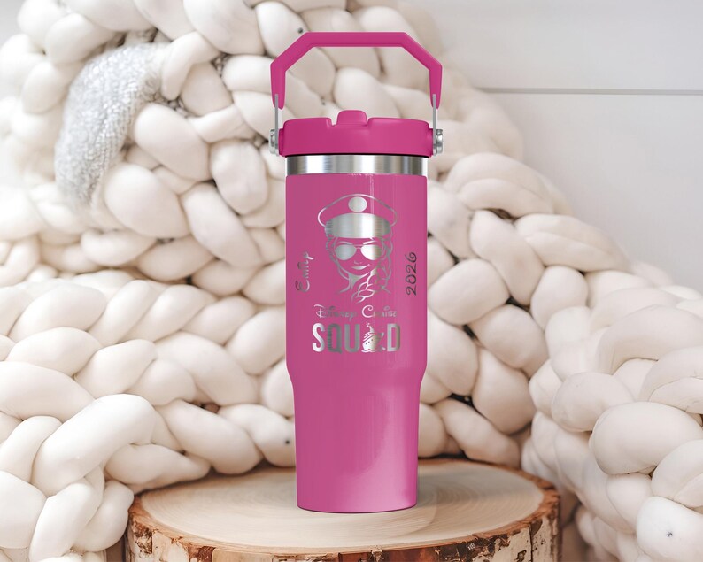 Puede incluir: Un vaso t&eacute;rmico rosa con asa y tapa a juego. El vaso presenta un gr&aacute;fico plateado de una mujer con gafas de sol y sombrero, con el texto "Squad" y el a&ntilde;o "2026". El vaso est&aacute; sobre una superficie de madera.