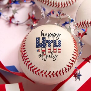 Könnte beinhalten: Ein weißer Baseball mit roten Nähten, der den Schriftzug Happy 4th of July in schwarzer Schrift und mit einem Sternen- und Streifenmuster gefüllten Buchstaben zeigt. Ein kleines Herzsymbol mit Flaggenmuster steht neben dem Text, umgeben von Sternen.