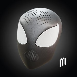 May include: A black, mask-shaped speaker with white mesh eye and mouth coverings. The speaker has a textured surface and small perforations on the top. A white logo is visible on the lower right.