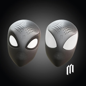 Puede incluir: Dos dispositivos de altavoz negros con forma de máscara de superhéroe, con formas de ojos blancos y pequeñas perforaciones en la parte superior. Un logotipo es visible en la parte inferior derecha. Los altavoces están sobre un fondo oscuro.