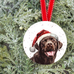 Pu&ograve; includere: Un addobbo natalizio rotondo con un Labrador Retriever marrone che indossa un cappello da Babbo Natale rosso. Il cane guarda direttamente lo spettatore con un'espressione felice. Lo sfondo &egrave; bianco con un effetto neve sottile.