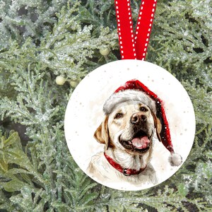 Pu&ograve; includere: Un ornamento natalizio in ceramica bianca con un'illustrazione ad acquerello di un labrador retriever giallo che indossa un cappello di Babbo Natale rosso. Il cane guarda lo spettatore con la lingua di fuori.