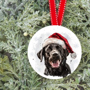 Pu&ograve; includere: Un Labrador Retriever nero che indossa un cappello rosso da Babbo Natale, su uno sfondo bianco innevato con fiocchi di neve. Il cane guarda direttamente lo spettatore con la bocca leggermente aperta, mostrando la lingua.