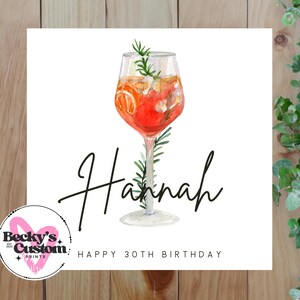 Op de afbeelding: Een vierkante witte kaart met een aquarel illustratie van een cocktail in een glas met voet. De naam "Hannah" staat in cursief geschreven, met "Happy 30th Birthday" eronder. De achtergrond is van hout.