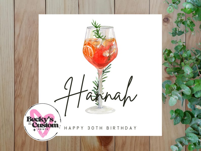 Op de afbeelding: Een vierkante witte kaart met een aquarel illustratie van een cocktail in een glas met voet. De naam "Hannah" staat in cursief geschreven, met "Happy 30th Birthday" eronder. De achtergrond is van hout.