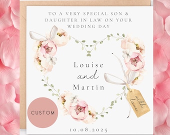 May include: A white card with a floral heart design and the text "To a very special son & daughter in law on your wedding day." The names "Louise and Martin" are written below the heart. The date "10.08.2025" is at the bottom of the card. A small tag with the text "with love" is hanging from the heart.