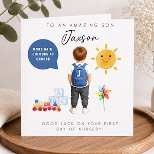 May include: A square greeting card featuring the text TO AN AMAZING SON Jaxson and GOOD LUCK ON YOUR FIRST DAY OF NURSERY. The design includes a child with a blue backpack, a yellow sun, a toy train with blocks, and a colourful pinwheel.