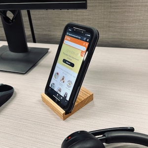 May include: A black smartphone with a wooden stand, displaying the eBay website with a sale banner that reads "Up to 50% off The Labor Day Sales Event is here!" The phone is on a light wood surface with a black headset.