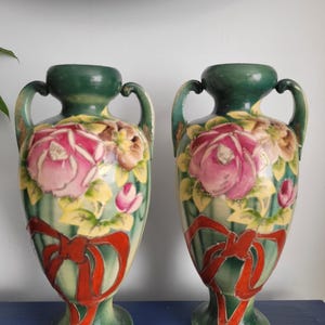 May include: Pair of antique ceramic vases. Each vase features a green base and handles, with a central design of pink roses and yellow leaves. A red ribbon design adorns the lower portion of the vases, adding a decorative touch. The vases are displayed on a blue surface.