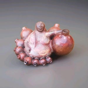 Puede incluir: Escultura de cerámica artesanal que representa una figura sentada rodeada de múltiples formas bulbosas. La escultura es principalmente de color marrón rojizo con un acabado liso y vidriado. La figura tiene una expresión serena y la pieza tiene un diseño único y orgánico.