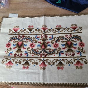 May include: A white embroidered throw pillow with a brown border and a floral design in shades of red, blue, and brown.