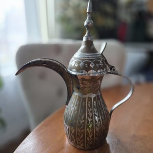 May include: An ornate, vintage brass coffee pot with silver accents. The pot features a long spout, a curved handle, and intricate floral and leaf patterns. The lid is topped with a decorative finial. The pot is sitting on a wooden table.