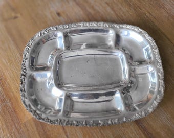 Pode incluir: Um prato de servir de prata com uma borda decorativa e cinco compartimentos. O prato tem forma oval e um acabamento brilhante.