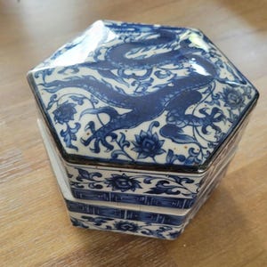 Pode incluir: Caixa hexagonal de cerâmica azul e branca com um desenho de dragão na tampa. A caixa tem um design em camadas com padrões florais e está colocada sobre uma superfície de madeira.