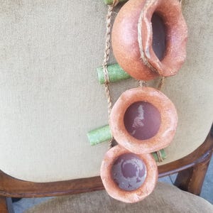 Könnte beinhalten: Handgefertigter, dekorativer Hängeornament mit drei terracottafarbenen, kreisförmigen Elementen. Die Elemente sind durch ein Naturseil und grüne zylindrische Teile verbunden. Das Ornament wird vor einem neutralen Hintergrund präsentiert.