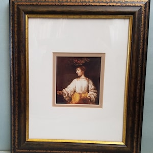 May include: A framed print of a painting depicting a woman wearing a white dress and a crown of flowers. The woman is holding a small object in her hand. The painting is set against a dark background.