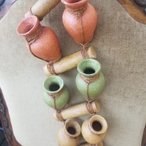 Puede incluir: Un adorno de pared decorativo con pequeños jarrones de cerámica en tonos naranja, verde y amarillo. Los jarrones están conectados por varillas de bambú de color beige y atados con hilo. La disposición se muestra sobre un fondo neutro.