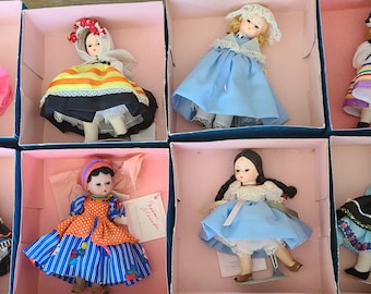 May include: Eight porcelain dolls in traditional dress from around the world, each in a separate pink box with a blue base. The dolls are wearing colorful dresses and hats, and some are holding accessories.