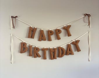 Könnte beinhalten: Ein braunes Filzbanner mit der Aufschrift "HAPPY BIRTHDAY" auf weißem Band, mit braunen Schleifen an jedem Ende. Die Buchstaben sind in einer fetten, abgerundeten Schriftart gehalten, und das Banner hängt an einer schlichten, hellen Wand. Eine festliche Dekoration für eine Geburtstagsfeier.