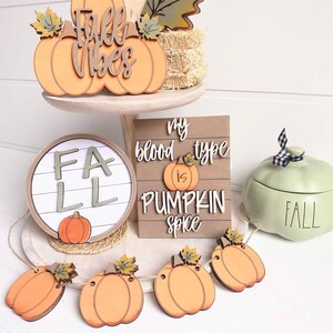 以下が含まれることがあります： 秋をテーマにした3段トレイの装飾。木製看板には「Fall Vibes」と刻まれ、かぼちゃと葉っぱが飾られています。丸い木製看板には「FALL」と刻まれ、木製看板には「My blood type is pumpkin spice」と刻まれています。かぼちゃのガーランドも飾られています。