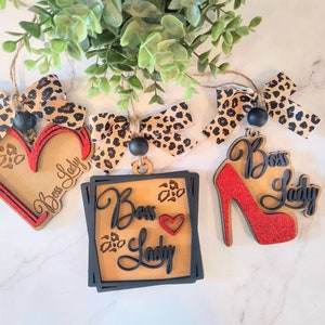 May include: Four wooden ornaments with a leopard print design and the text "Boss Lady". One ornament is a heart shape, one is a square, one is a high heel shoe, and one is a bow.