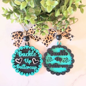 Puede incluir: Dos adornos de madera azul turquesa y negro con el texto "Buckle Up Buttercup" y un diseño de corazón. Cada adorno tiene una cinta de estampado de leopardo.