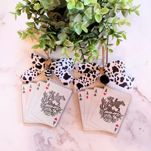May include: Wooden playing card ornaments featuring an ace of spades design with a cowboy on a horse, roses, and cacti. The ornaments are paired with black and white cow print bows and a green plant.