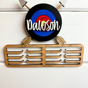 Puede incluir: Porta medallas de madera para tiro con arco con un blanco negro y rojo y el nombre "Dawson" en blanco. El porta medallas tiene ocho ganchos para medallas.