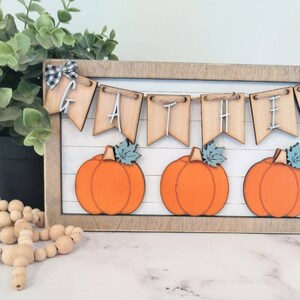 May include: A wooden framed sign with a banner that reads "GATHER" and three orange pumpkins. The banner is made of wood and has a rustic look. The pumpkins are painted orange and have green stems and leaves.