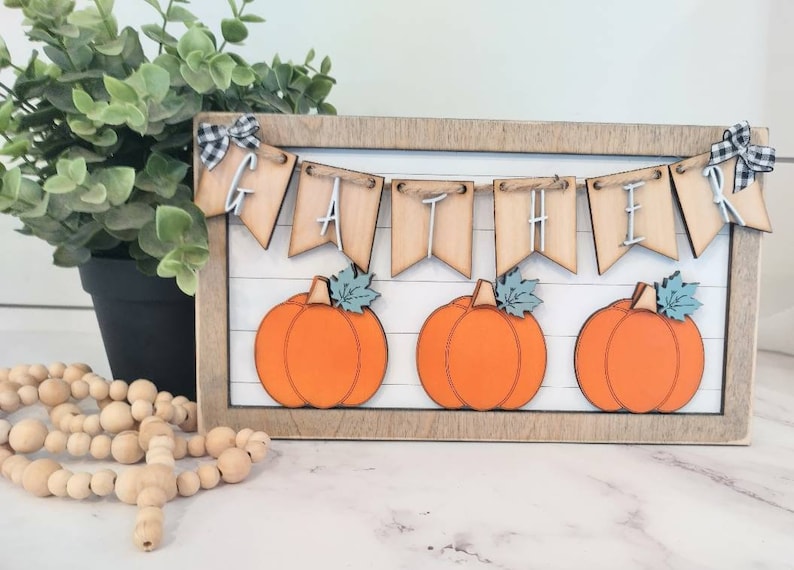 May include: A wooden framed sign with a banner that reads "GATHER" and three orange pumpkins. The banner is made of wood and has a rustic look. The pumpkins are painted orange and have green stems and leaves.