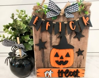 Puede incluir: Un letrero de madera con una calabaza naranja y negra, estrellas negras y una pancarta que dice "Trick or Treat". El letrero está colgado de una cuerda y tiene una cinta a cuadros negros y blancos.