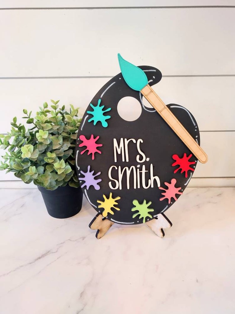 Op de afbeelding: Een zwart schoolbord in de vorm van een palet met een bevestigd houten kwast. Het palet heeft kleurrijke verfspatten en de tekst "Mrs. Smith" erop geschreven. Het palet staat op een houten standaard.