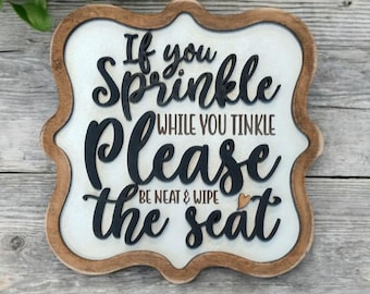 May include: Wooden bathroom sign with a white background and brown trim. The sign has the text "If you sprinkle while you tinkle please be neat & wipe the seat" in black lettering.