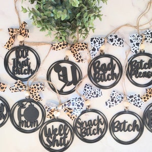 May include: A set of 12 black wooden ornaments with various phrases and designs, including "100% That Bitch", "Bad Bitch", "Bitch Please", "Crazy Bitch", "Boss Bitch", "Snack Bitch", "Well Shit", and "Mom". The ornaments are strung together with twine and have leopard print bows.