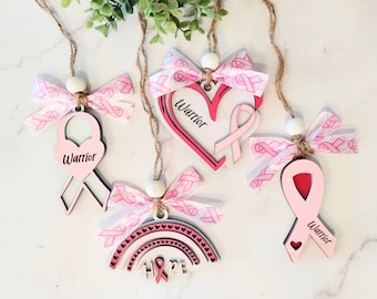 Könnte beinhalten: Vier rosafarbene und weiße Holzornamente mit rosa Bändern und dem Wort "Warrior" auf jedem. Ein Ornament ist herzförmig, eines ist bandförmig, eines ist bandförmig mit einem Herz in der Mitte und eines ist regenbogenförmig mit dem Wort "Hope" in der Mitte.