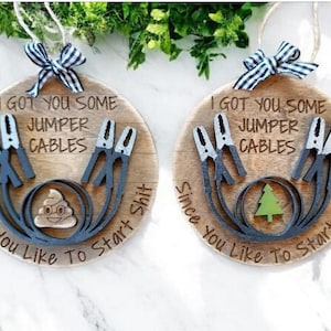 Op de afbeelding: Twee ronde houten ornamenten met de tekst "I GOT YOU SOME JUMPER CABLES Since You Like To Start Shit". Elk ornament heeft startkabels en een afbeelding van een lachende poep-emoji of een groene kerstboom.