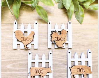 May include: Four wooden ornaments shaped like picket fences with farm animal silhouettes inside. The animals are a chicken, a cow, a pig, and another chicken. The ornaments are white with brown animal silhouettes and the words "cluck", "moo", "oink", and "cluck" are engraved on the animals.