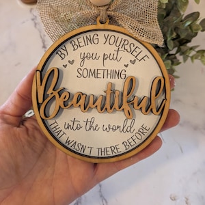 May include: A round wooden ornament featuring the text BY BEING YOURSELF you put SOMETHING Beautiful INTO THE WORLD THAT WASN'T THERE BEFORE. The piece includes a natural wood bead, a burlap ribbon bow, and a jute twine hanger for display.