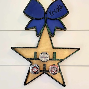 May include: A wooden star-shaped ring holder with green glitter trim. The star has three slots for rings. There is a blue bow with green glitter trim and the name "Krista" written on it above the star.