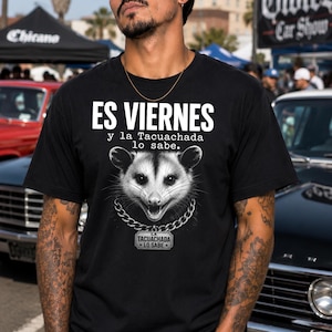 May include: A black short-sleeved t-shirt featuring a graphic of an opossum wearing a silver chain with a rectangular tag. The text reads ES VIERNES y la Tacuachada lo sabe. The tag on the chain also displays the text LA TACUACHADA LO SABE.